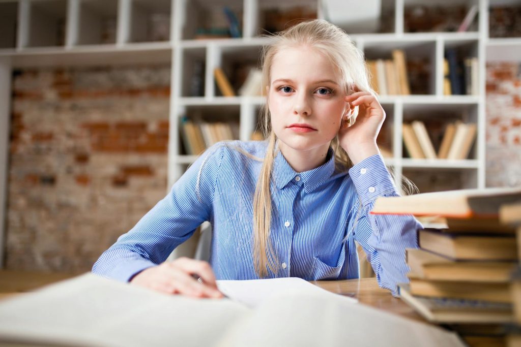 A young woman with blond hair studying at a table piled with books.