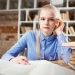 A young woman with blond hair studying at a table piled with books.