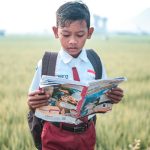 A young schoolboy in uniform reads a magazine in a grassy field, emphasizing education.