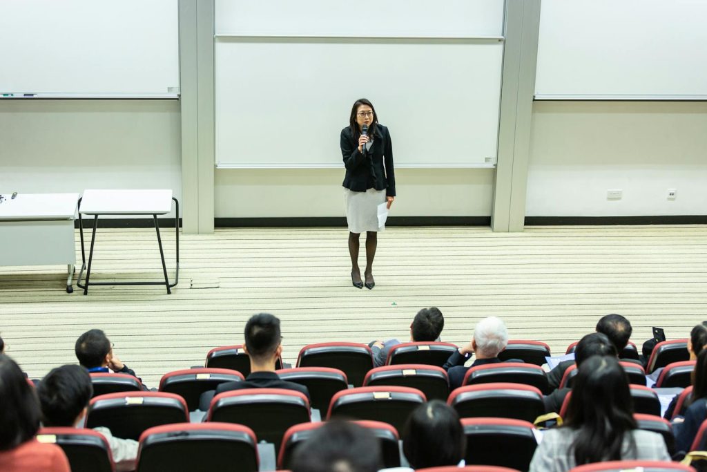 Female speaker presenting to an audience in a modern auditorium setting.