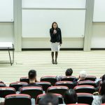 Female speaker presenting to an audience in a modern auditorium setting.