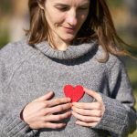 A woman outdoors holding a red heart against her chest, capturing a warm moment.