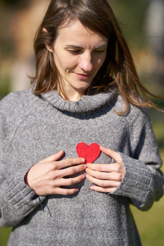 A woman outdoors holding a red heart against her chest, capturing a warm moment.