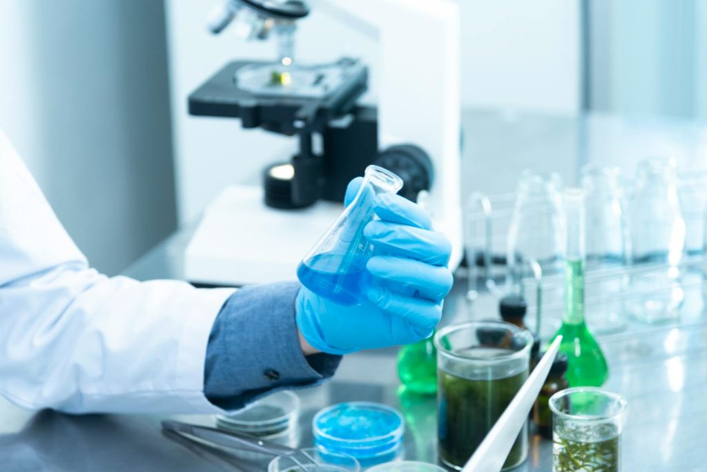 Scientist in gloves analyzing blue liquid in a laboratory setting with microscope and glassware.