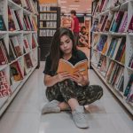 Young woman sitting on the floor and reading a book in a modern bookstore with shelves full of colorful books.