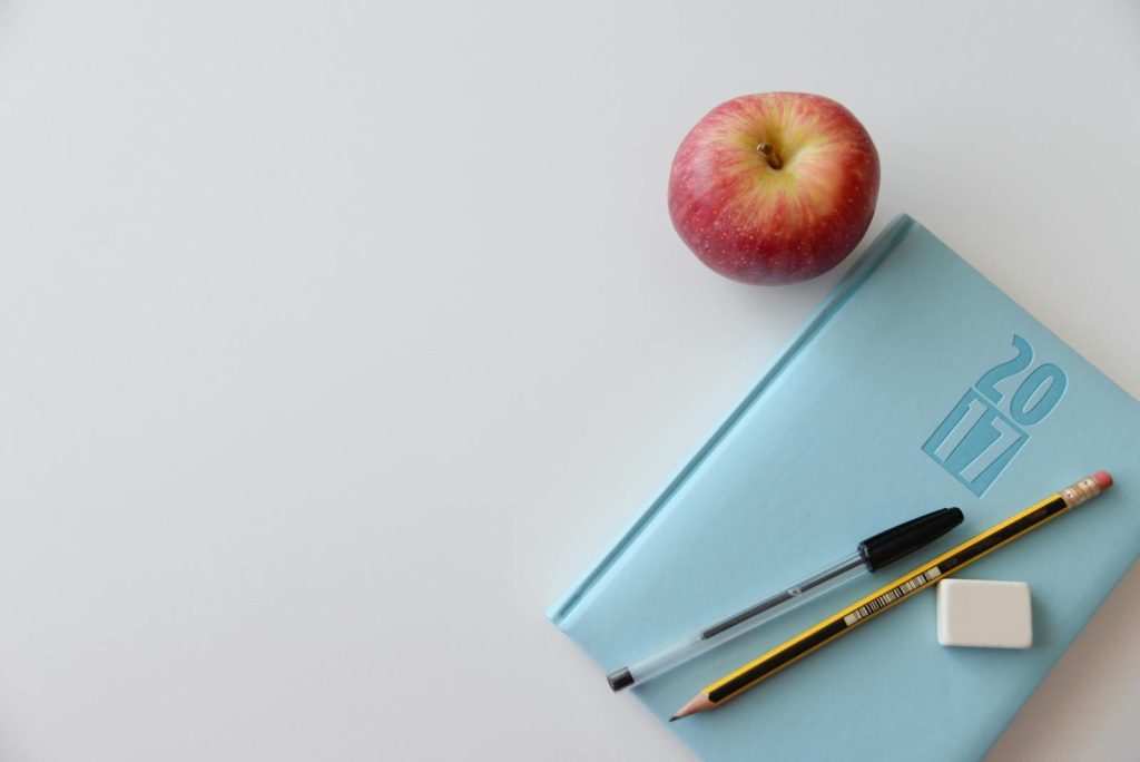 A minimalist flat lay of a study desk with a red apple, blue journal, pencil, pen, and eraser.