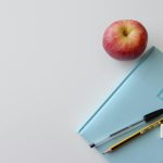 A minimalist flat lay of a study desk with a red apple, blue journal, pencil, pen, and eraser.