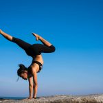 A woman performing a yoga handstand on a rock by the sea under a clear blue sky.