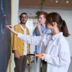 A group of young people discussing ideas at a blackboard in a modern office setting.