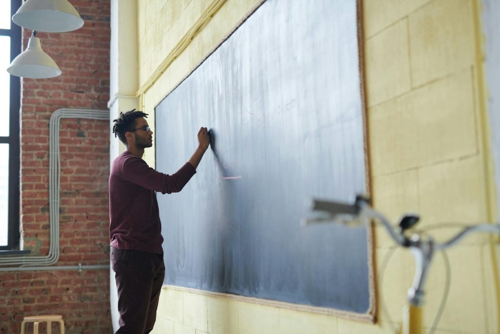 Man with dreadlocks writing on a blackboard in a classroom setting.