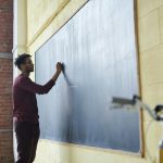 Man with dreadlocks writing on a blackboard in a classroom setting.