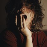 A woman with bed hair yawns in dim lighting, depicting fatigue and morning mood.