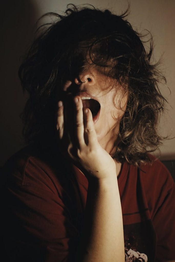 A woman with bed hair yawns in dim lighting, depicting fatigue and morning mood.