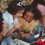 A group of happy young women enjoying a sunny picnic outdoors, engaging with a smartphone and laughing.