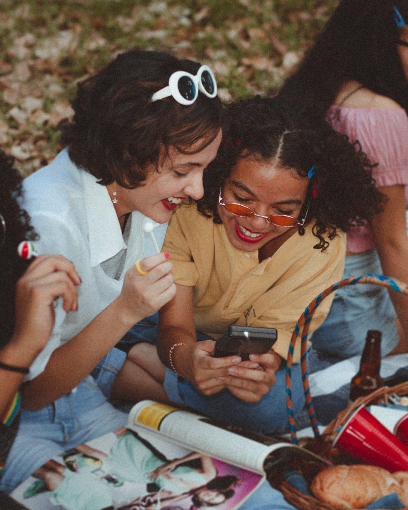 A group of happy young women enjoying a sunny picnic outdoors, engaging with a smartphone and laughing.