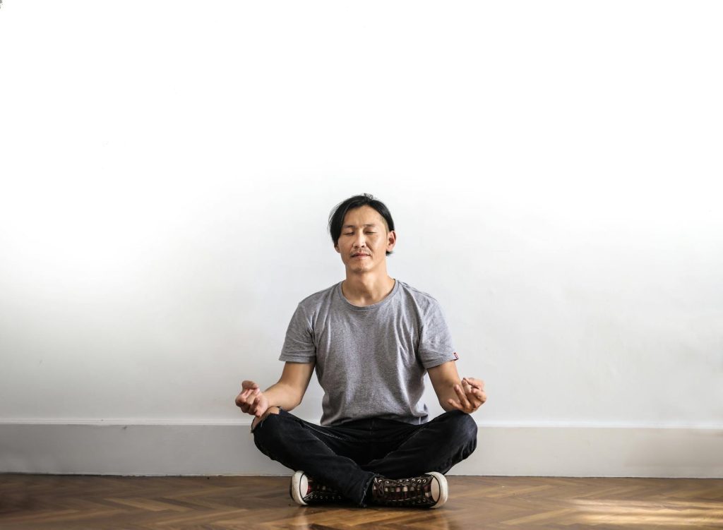 Asian man practicing meditation indoors on a wooden floor, promoting relaxation and mindfulness.