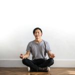 Asian man practicing meditation indoors on a wooden floor, promoting relaxation and mindfulness.