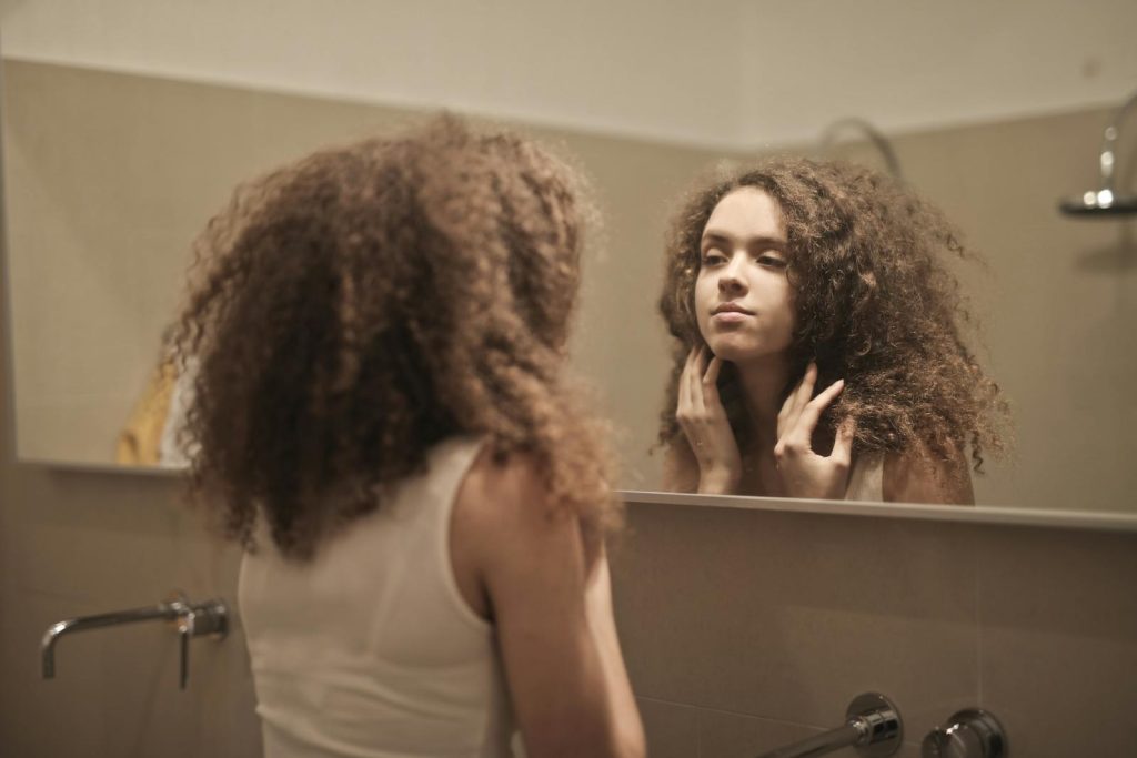 A woman with curly hair contemplates in front of a bathroom mirror, reflecting on her thoughts.