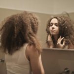 A woman with curly hair contemplates in front of a bathroom mirror, reflecting on her thoughts.