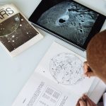 Crop of faceless astronomer in uniform modifying astronomical map while sitting at table with tablet and educational materials conducting research at science center