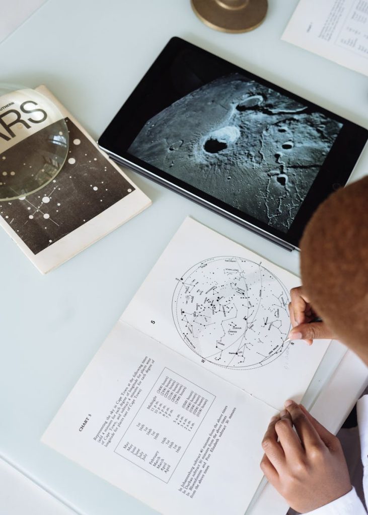 Crop of faceless astronomer in uniform modifying astronomical map while sitting at table with tablet and educational materials conducting research at science center