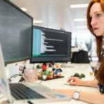 A focused female software engineer coding on dual monitors in a modern office.