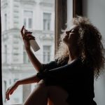 Relaxed young woman with curly hair sprays hydrating mist by a window sill in urban setting.