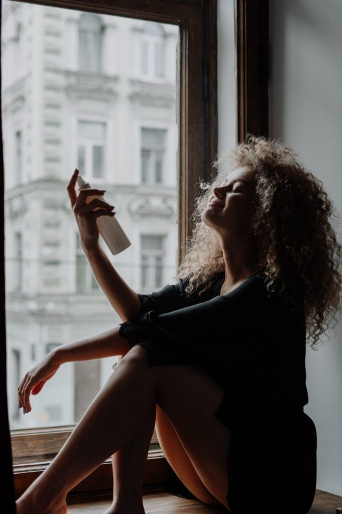 Relaxed young woman with curly hair sprays hydrating mist by a window sill in urban setting.