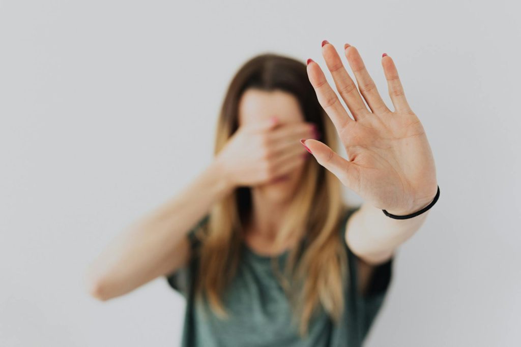 A woman in a gray shirt covers her face with her hand in a stop gesture, evoking a sense of fear and protection.