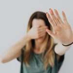 A woman in a gray shirt covers her face with her hand in a stop gesture, evoking a sense of fear and protection.