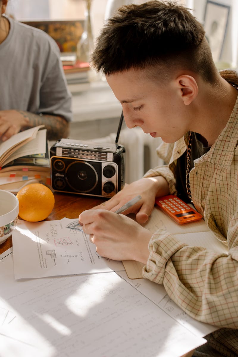 Young student studying with textbooks, calculator, and radio on a sunny day indoors.