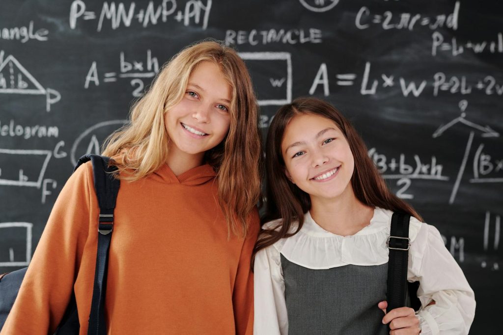 Two cheerful girls stand in a classroom with a math-covered chalkboard, ready for learning.