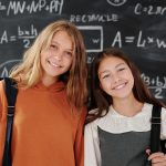 Two cheerful girls stand in a classroom with a math-covered chalkboard, ready for learning.