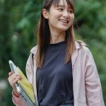 Smiling young Asian woman carrying a laptop and books, enjoying a sunny day outdoors.