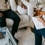 From above crop African American male in casual clothes sitting on comfy couch and answering professional female consultant questions in modern psychotherapy office