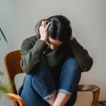 A young woman sitting on a chair indoors, holding her head in distress.