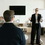 Businesswoman giving presentation in office with bookshelves and clock.