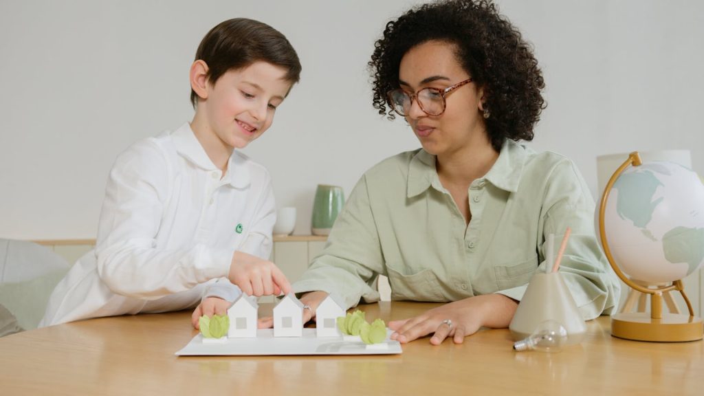 A young boy and a teacher exploring sustainable energy models with model houses and a globe in a classroom setting.