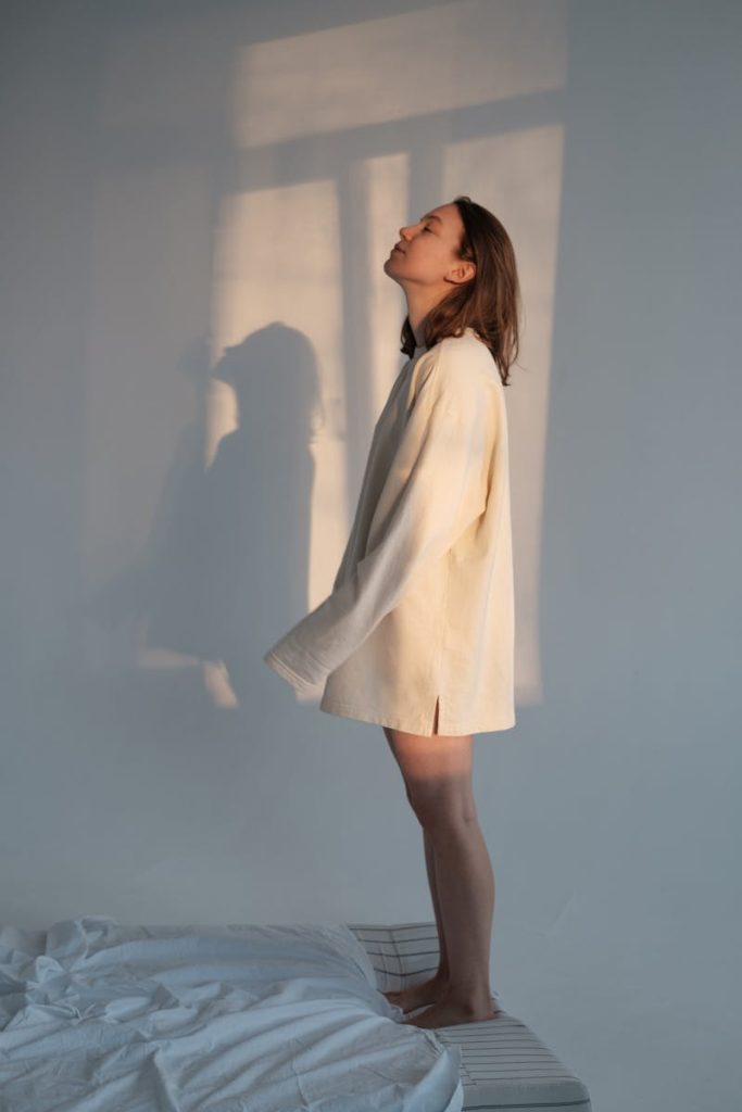 A serene young woman stands indoors, bathed in sunlight, wearing a casual sweater.