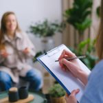 Unrecognizable professional female psychologist writing on clipboard while sitting against client on blurred background during psychotherapy session in light office