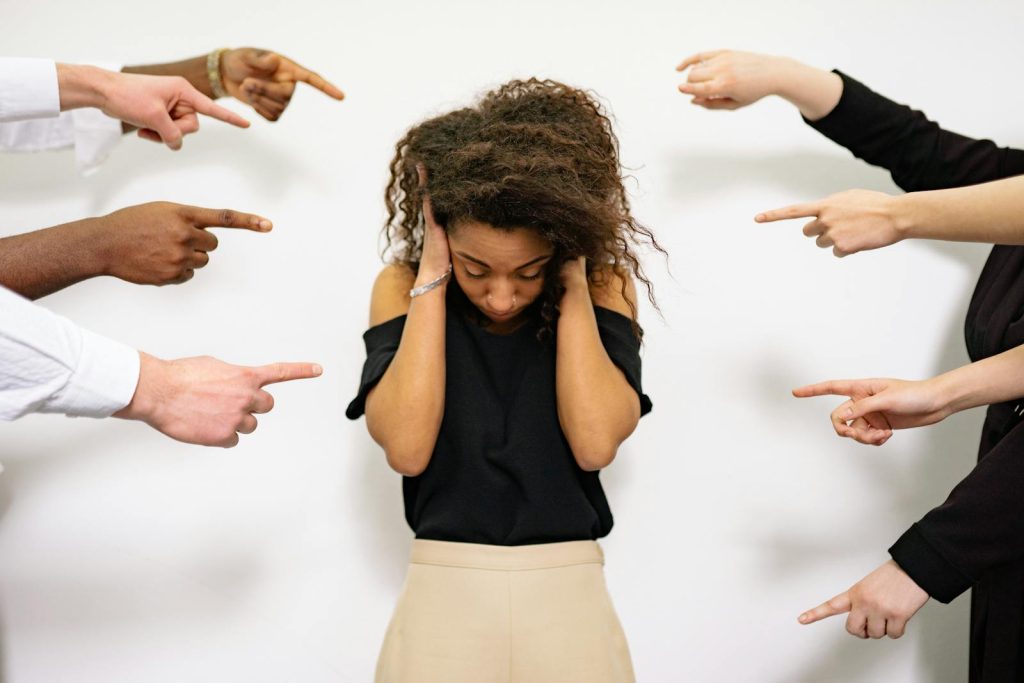 A young woman with curly hair looks distressed as multiple hands point accusatory fingers at her.