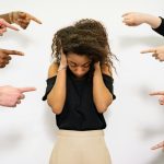 A young woman with curly hair looks distressed as multiple hands point accusatory fingers at her.