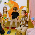 Young children playing musical bells in a colorful classroom setting with a teacher.