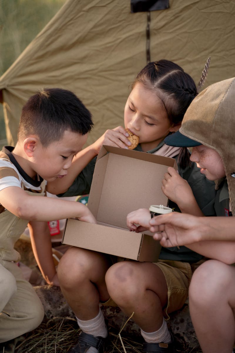 Three children sharing snacks outdoors at a summer scouting camp.