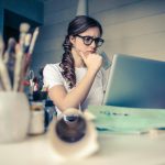 Young woman with glasses deeply focused on a laptop surrounded by art supplies in a home office.